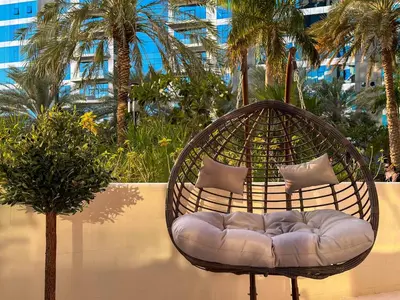a rattan chair with pillows sitting next to a tree at Upgraded 1BR Paradise With Private Garden - Oceana in Dubai