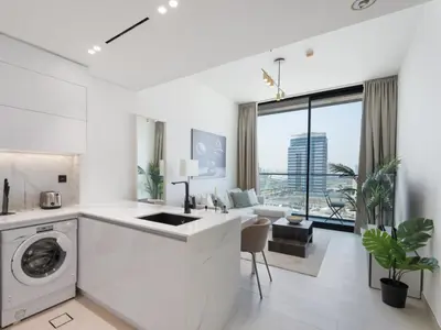 a white kitchen with a washing machine and a living room at Luxe Escape - Stylish 1BR in Binghatti Venus-05 in Dubai
