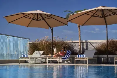 a woman sitting on a table next to a pool with umbrellas at Vogue JVT Novotel near Springs Souk Mall, One Bedroom apartment with Balcony and Vibrant Community View, Access to all Amenities in Dubai