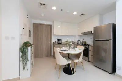 a white kitchen with a table and a refrigerator at BNew Chic 1BR Apt Near Trump Golf w Pool and Gym in Dubai